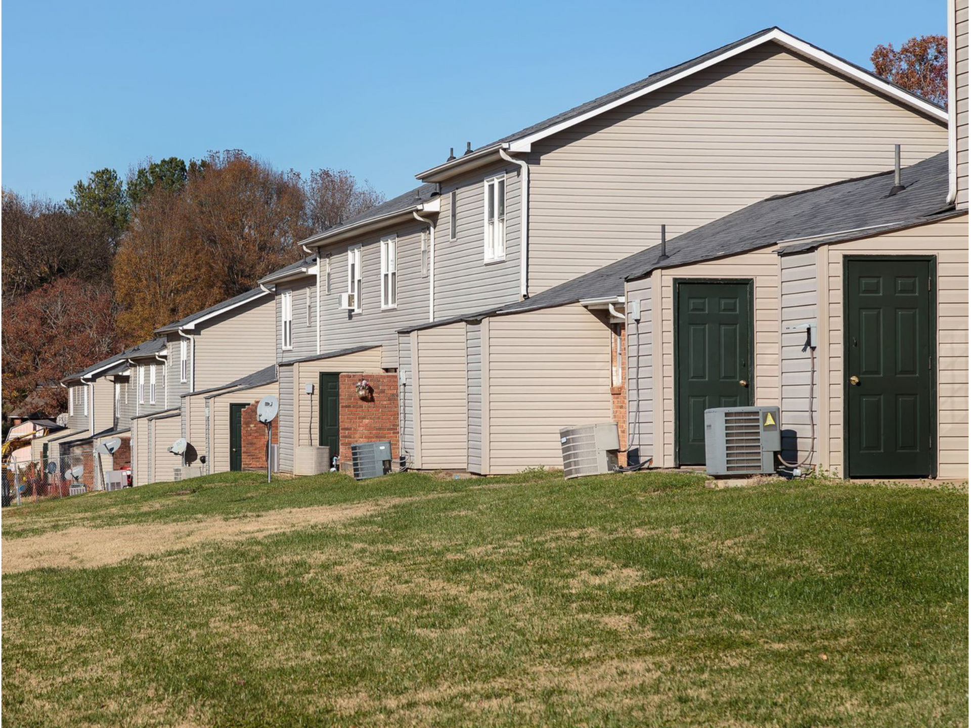 Row of beige townhouse-style apartment buildings with green doors and outdoor AC units along a grassy lawn.