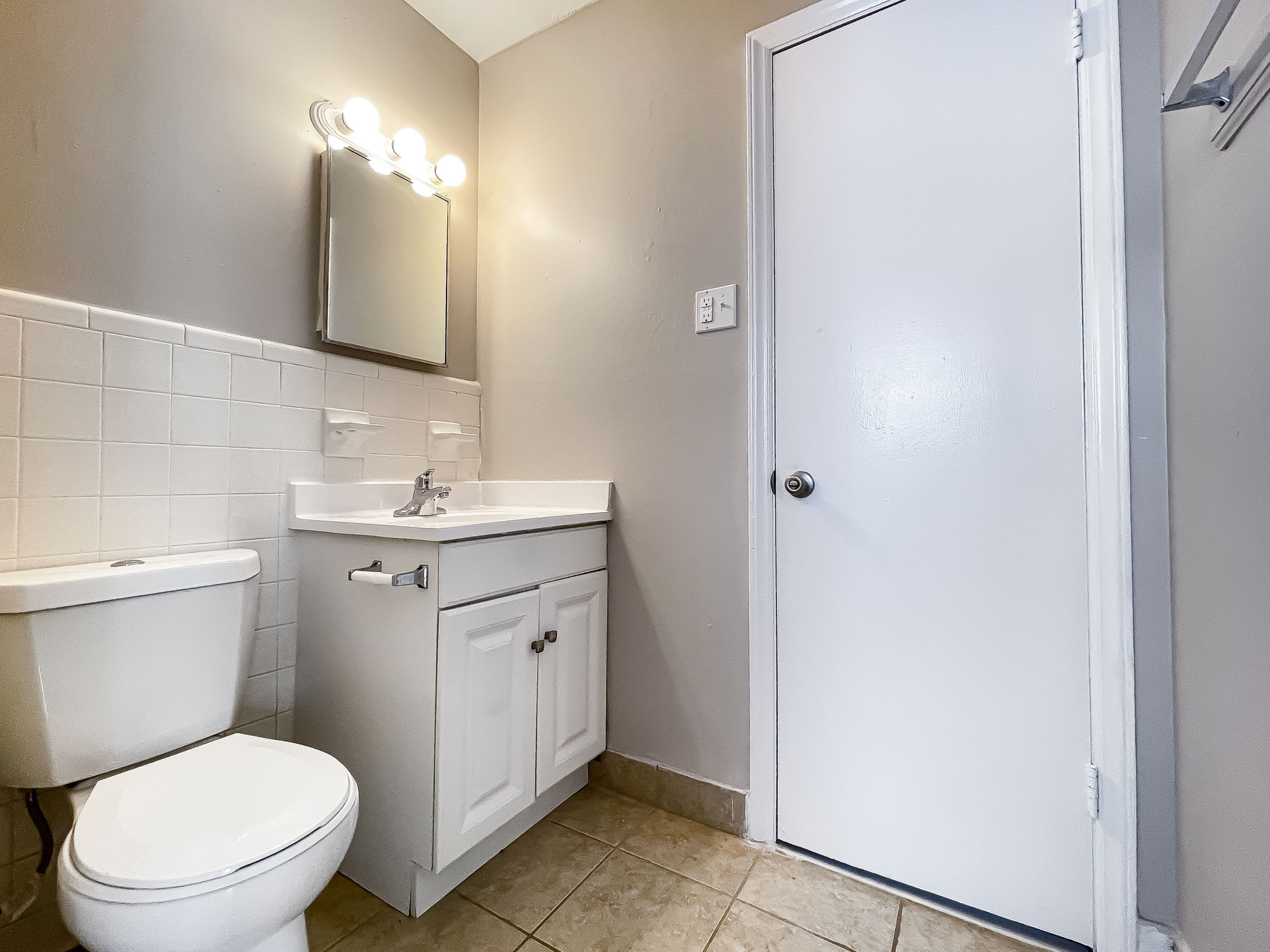 Bathroom with white vanity, toilet, mirror with bulbs, and a closed door.