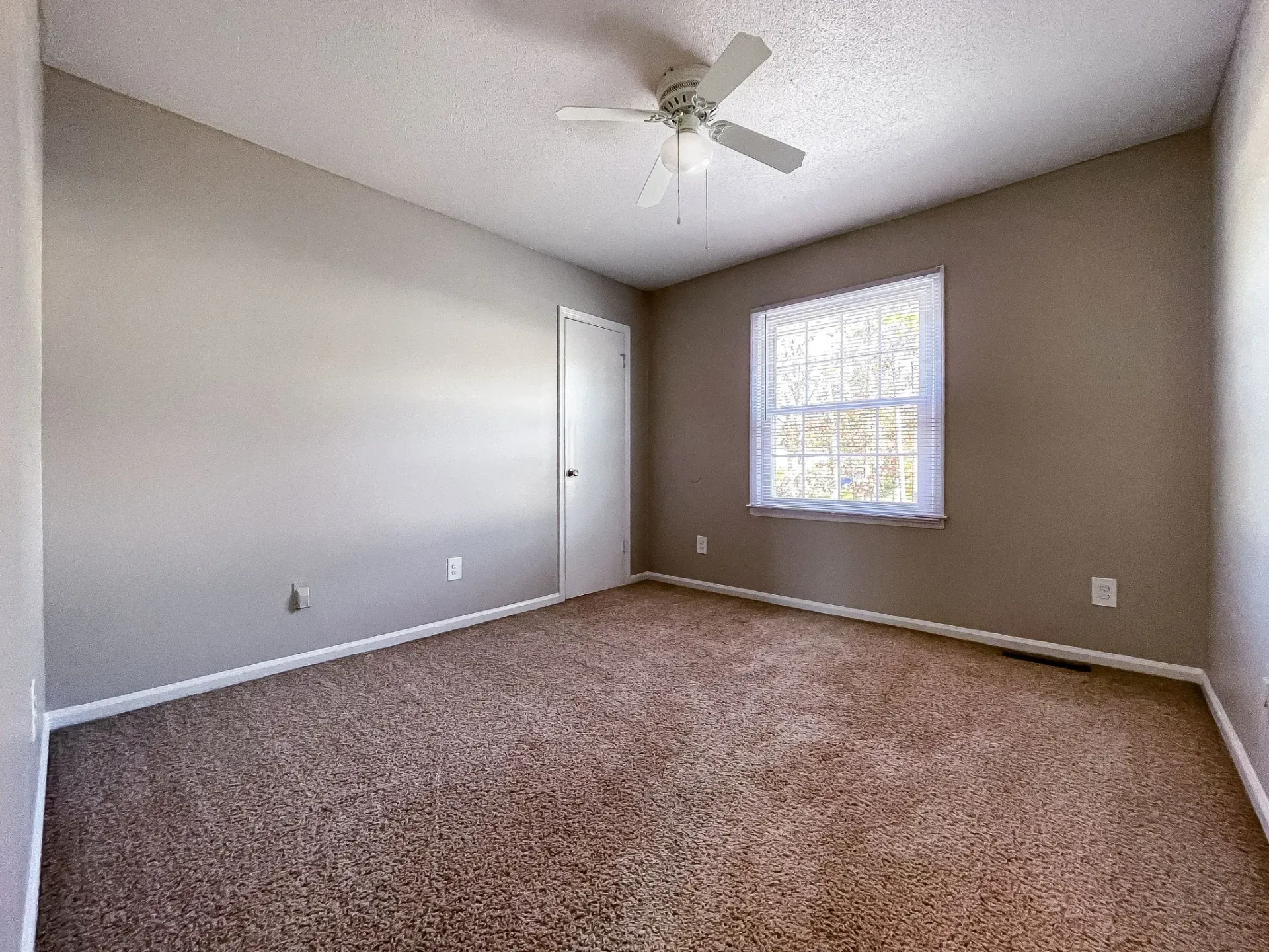 Empty apartment bedroom with beige walls, carpet, a window, and a ceiling fan.