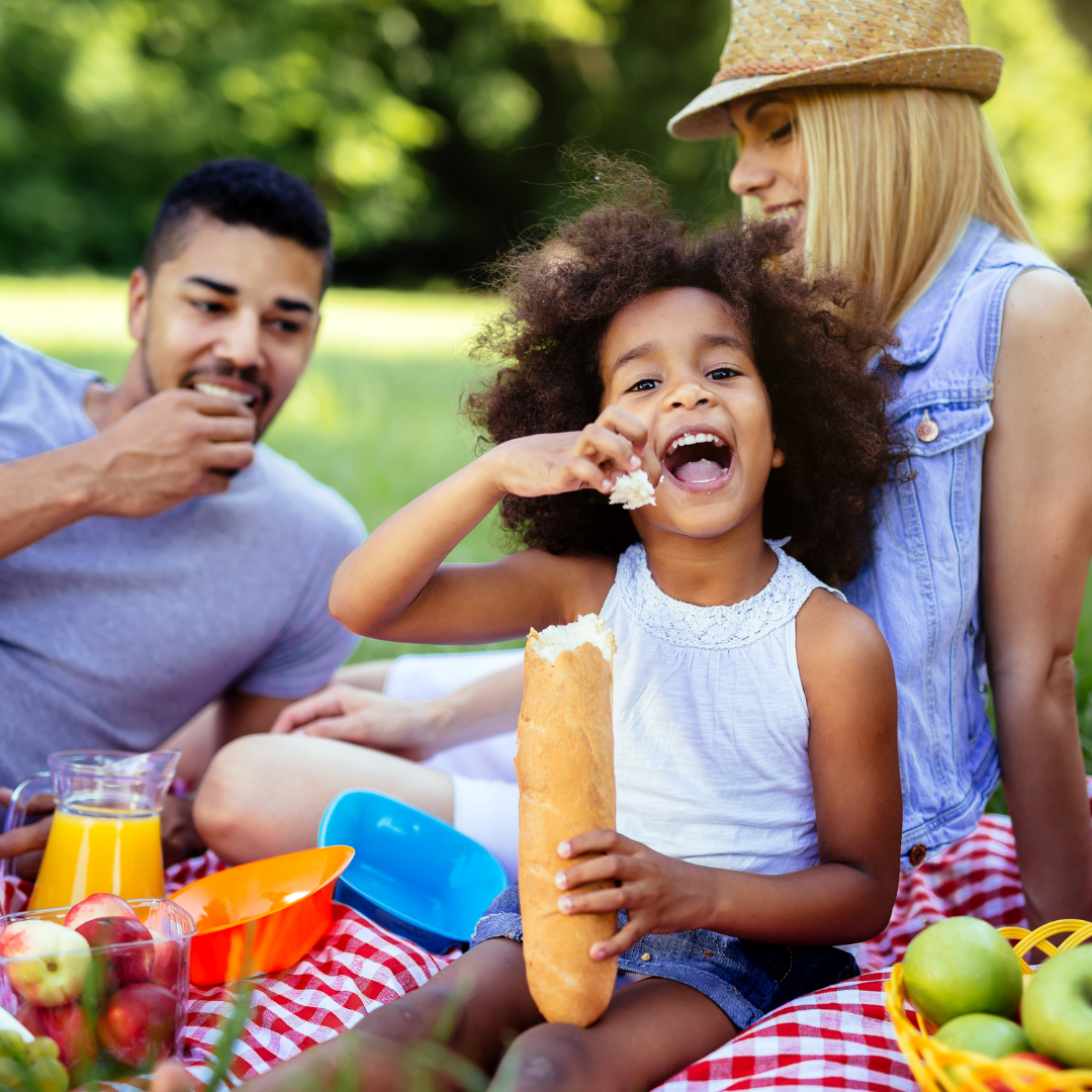 Family picnic: laughing Black girl, eating bread, with parents on checkered blanket. Outdoors.
