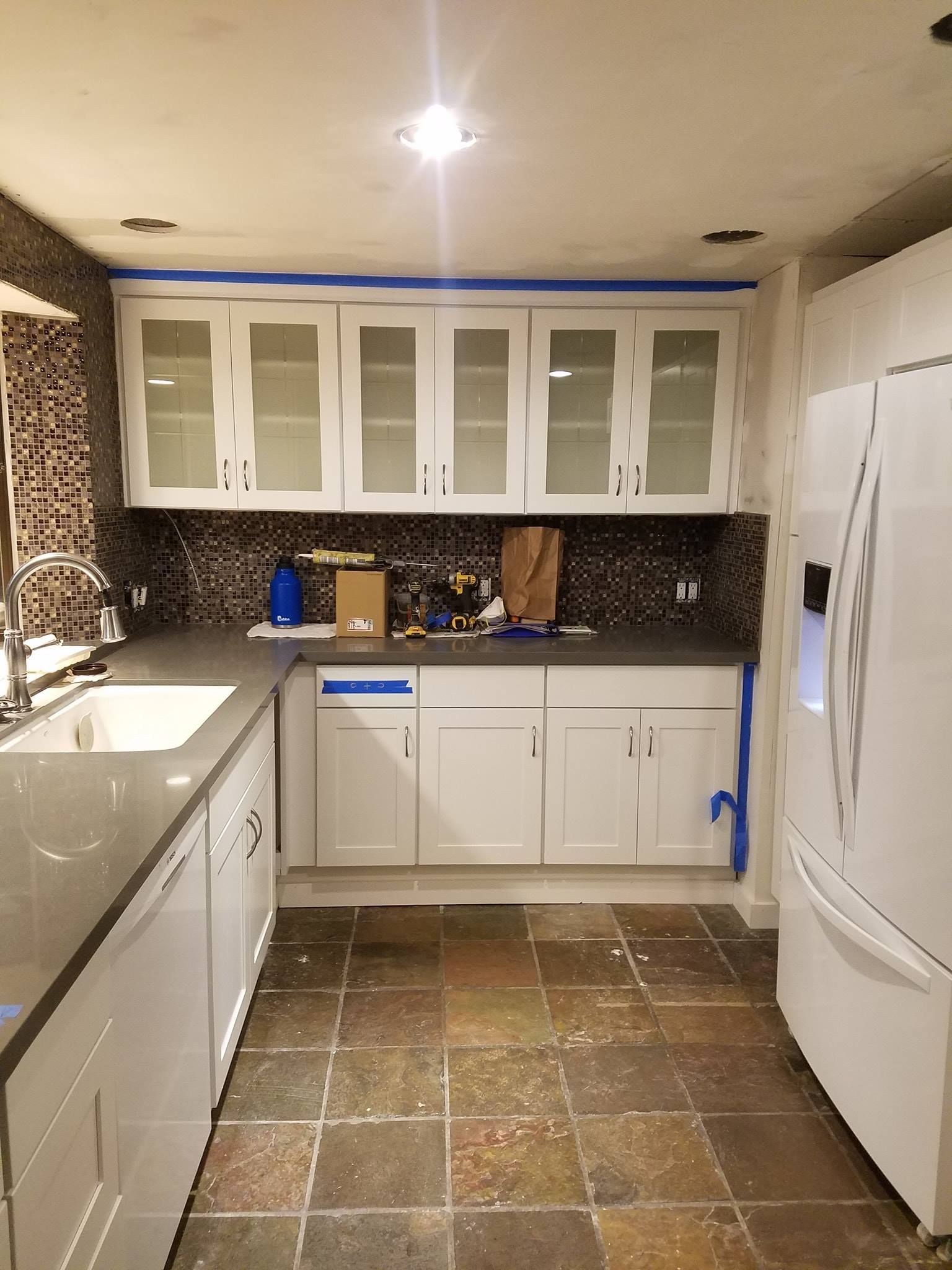Kitchen with white cabinets, gray countertops, and a brown tiled floor, mid-renovation.
