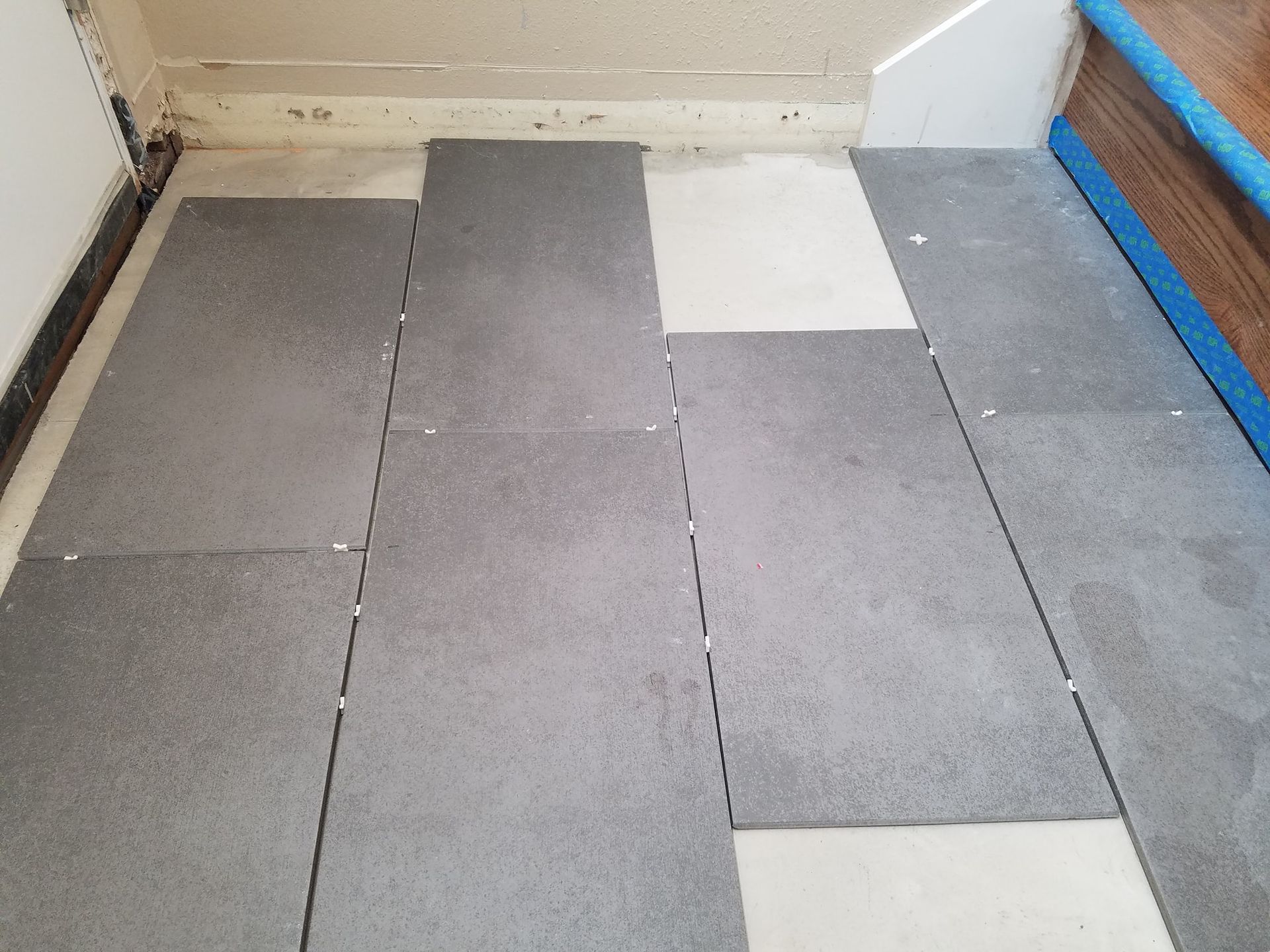 Gray floor tiles in a staggered pattern on a light-colored surface near a door and a stair rail.
