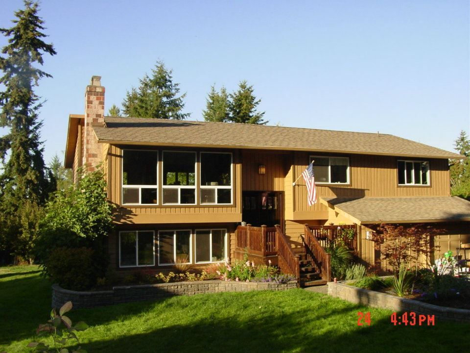 Two-story brown house with large windows, wooden deck, and American flag; sunny day.