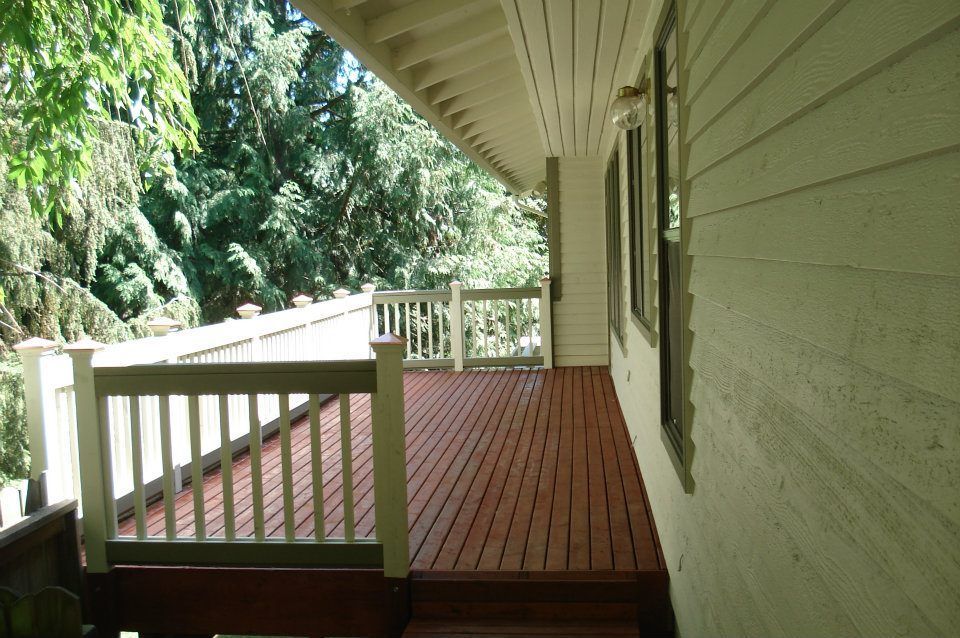Deck with brown wood and white railings, against a house with green trim, surrounded by trees.