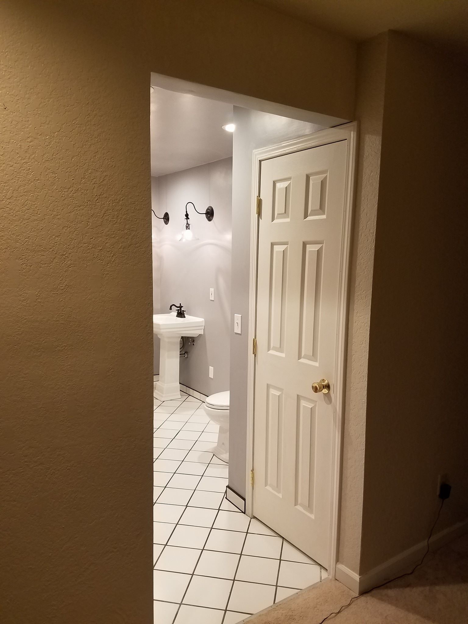 Open white door leading into a bathroom with white pedestal sink and black accents on white tiled floor.