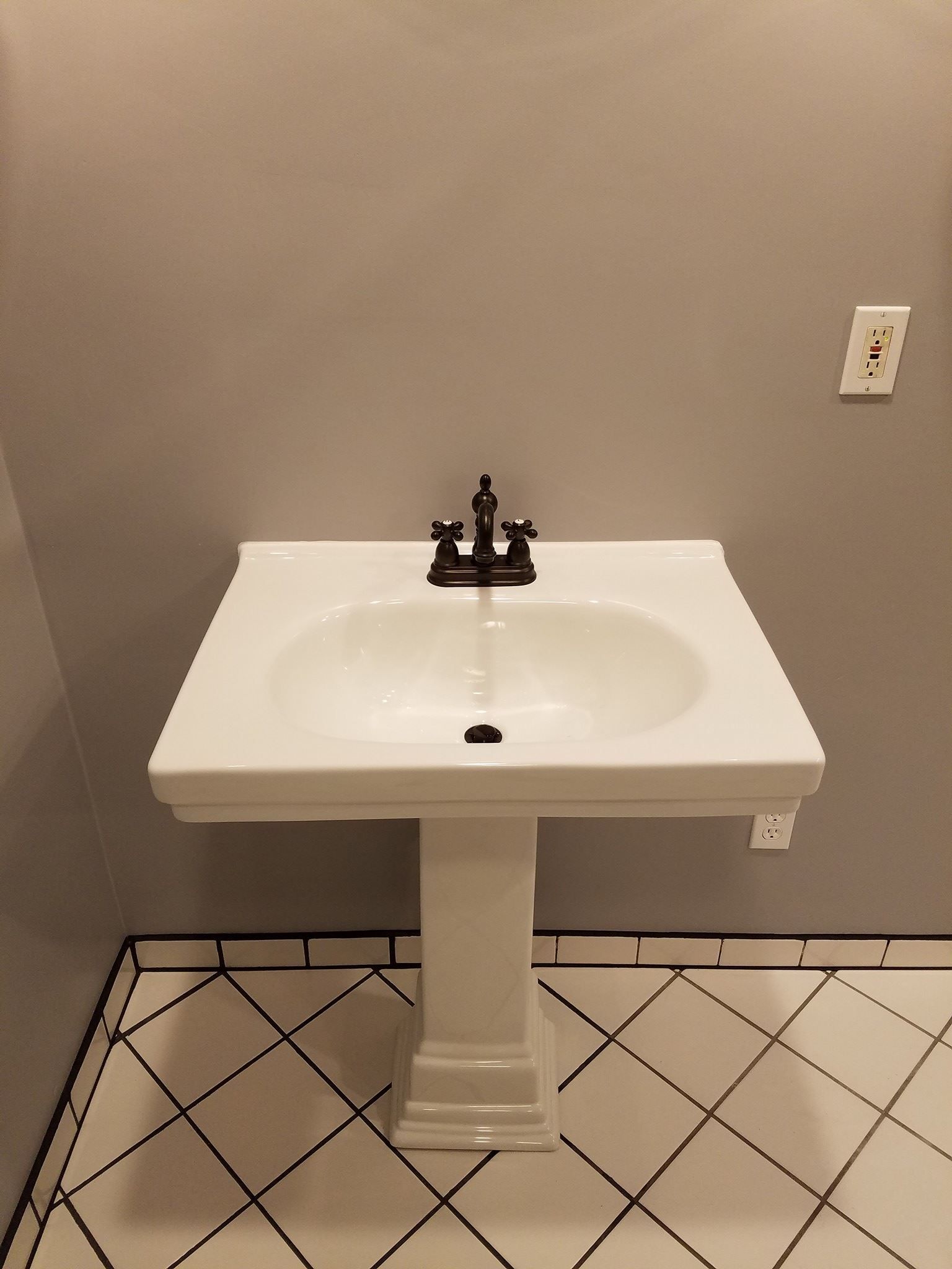 White pedestal sink with black fixtures and a diamond-patterned tile floor against a grey wall.