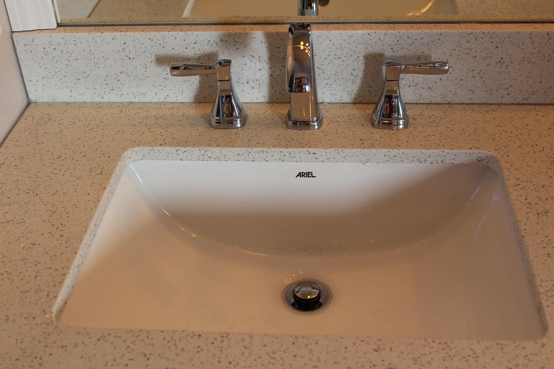 White bathroom sink with chrome faucet and handles on speckled countertop.
