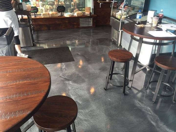 Interior of a cafe with round tables, stools, and metallic floor. A person stands near a counter with food.