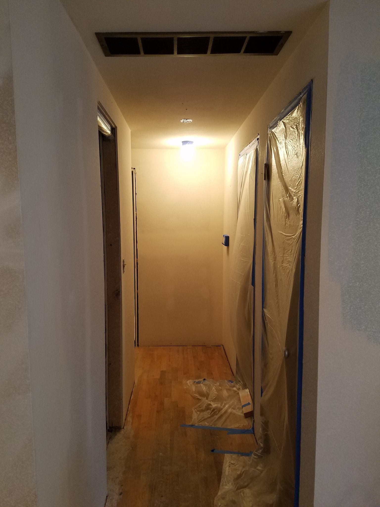 Narrow hallway with wood floor and white walls, two doorways, and one door covered in plastic.