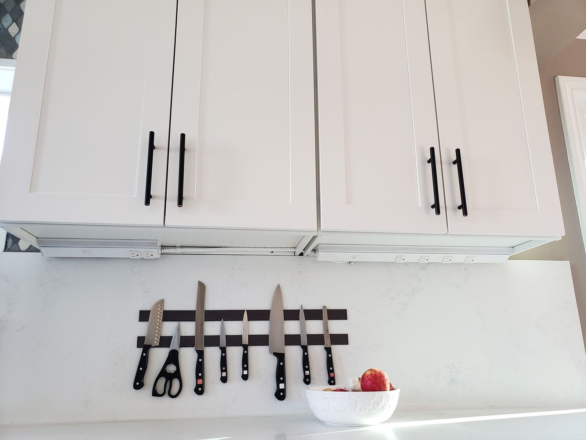 White kitchen cabinets with black handles, above a magnetic knife holder. A bowl of apples sits on the counter.