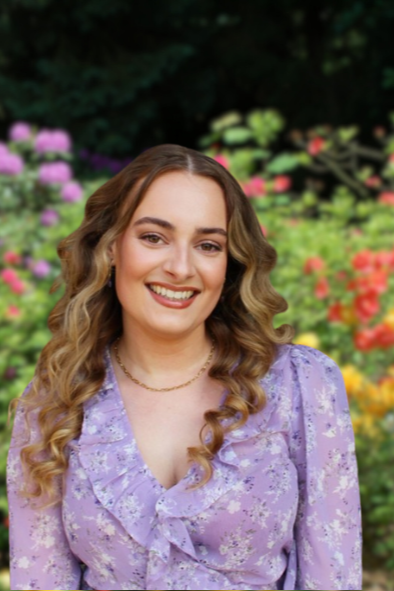 Een vrouw in een pak staat in een tuin, met haar handen gevouwen en glimlachend.