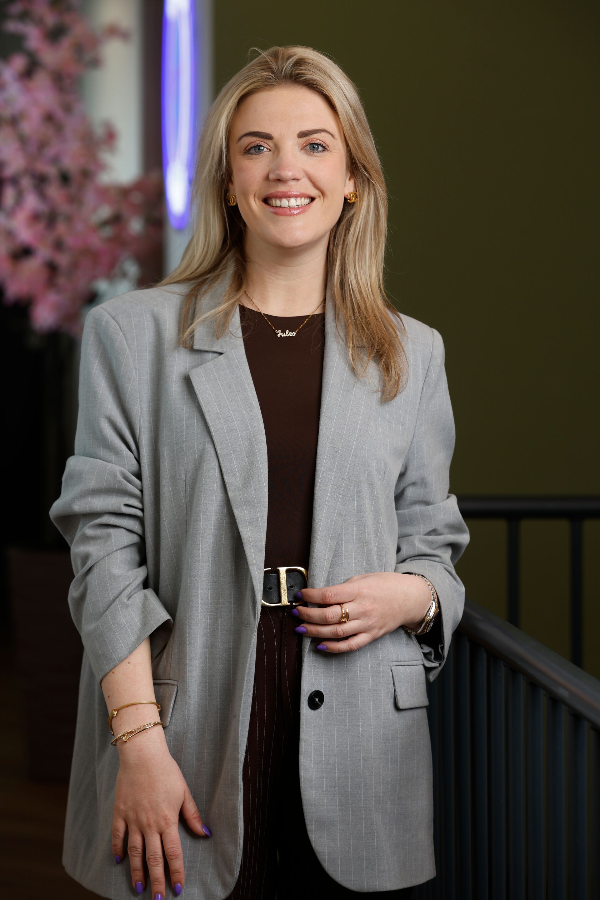 Een vrouw met een hand ik haar zak, ze staat voor de heg en lacht naar de camera