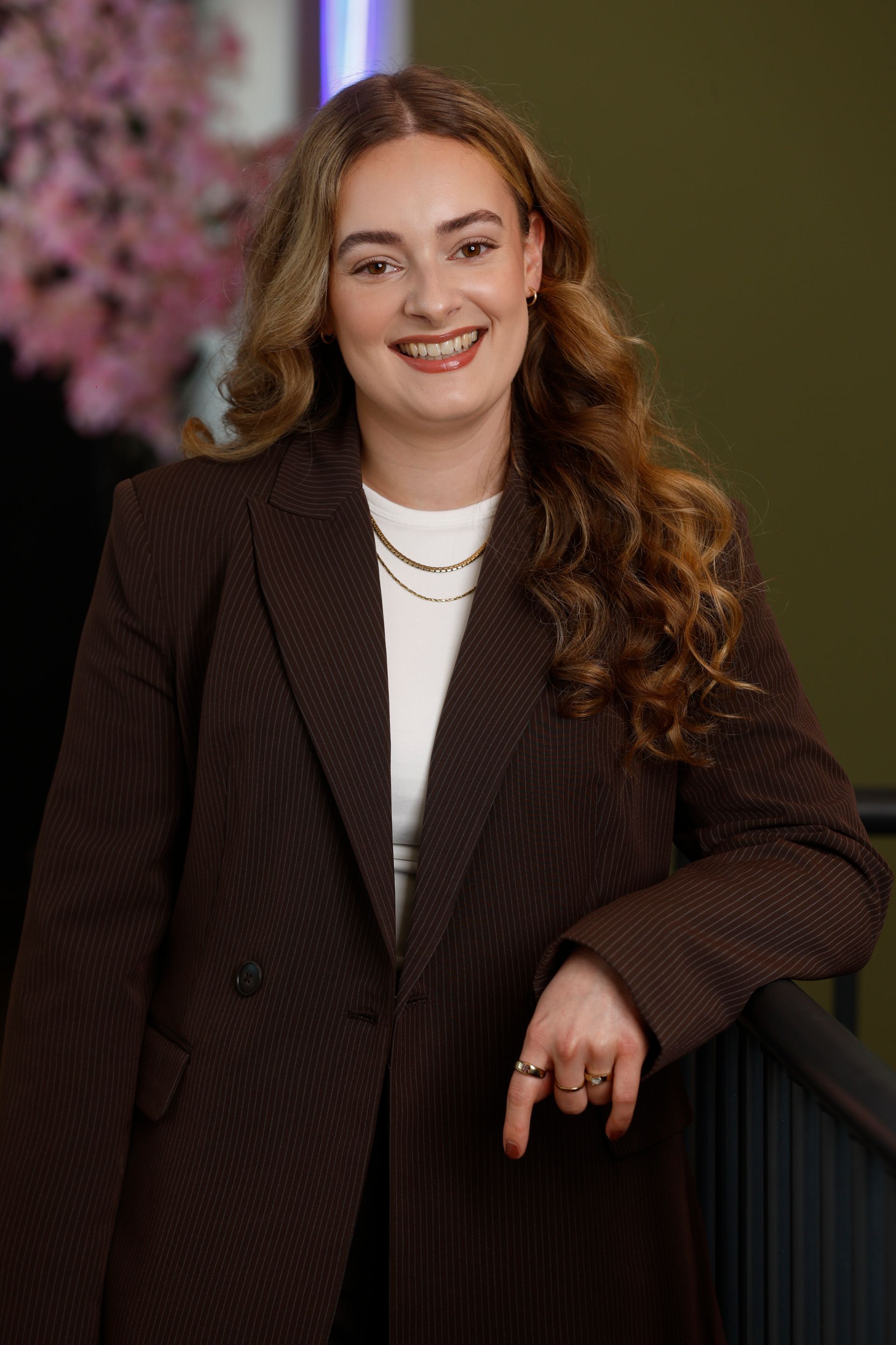 Een vrouw in een pak staat in een tuin, met haar handen gevouwen en glimlachend.