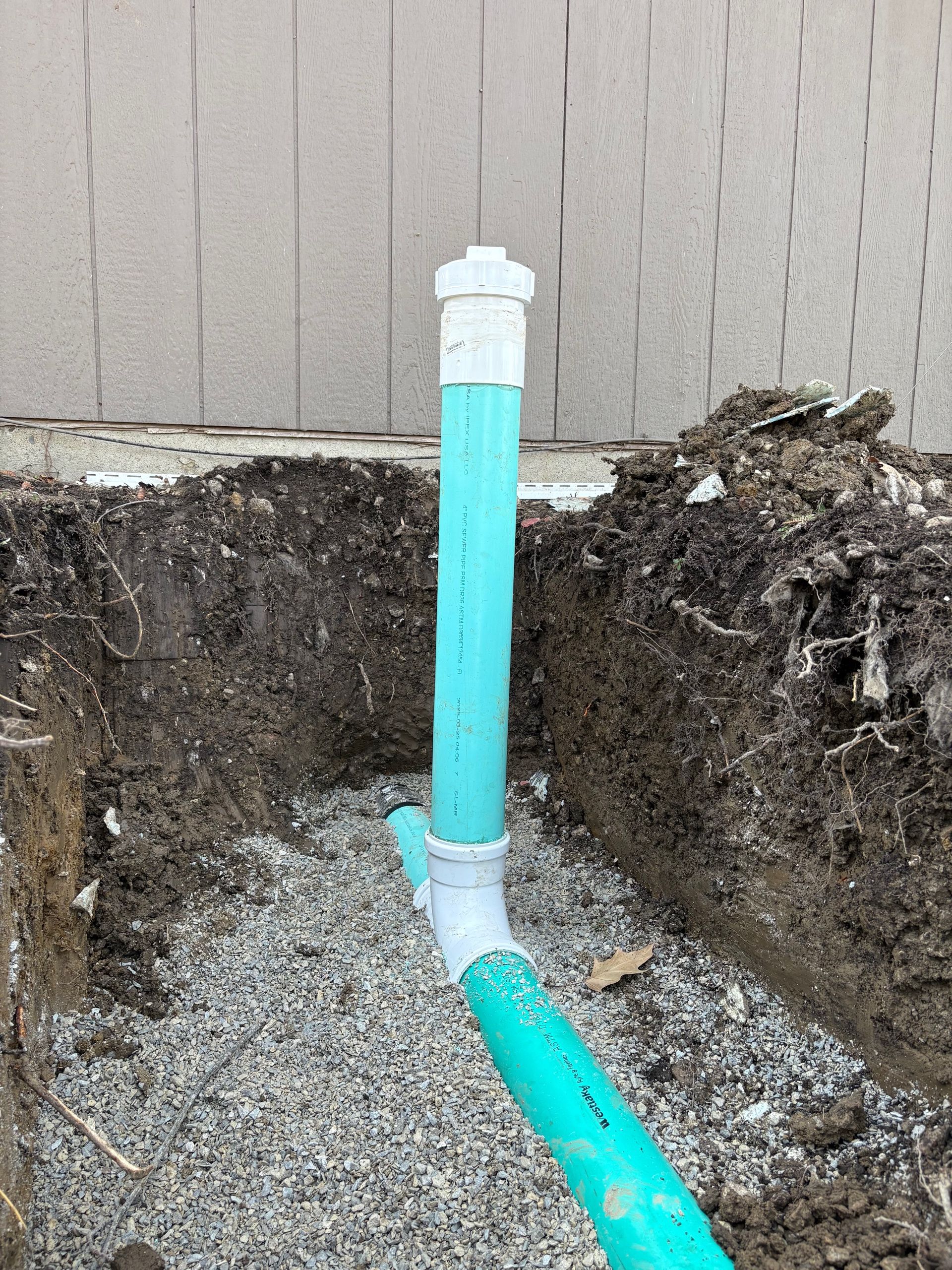 Turquoise PVC pipe with a white cap rising from a dug-out trench beside a wall