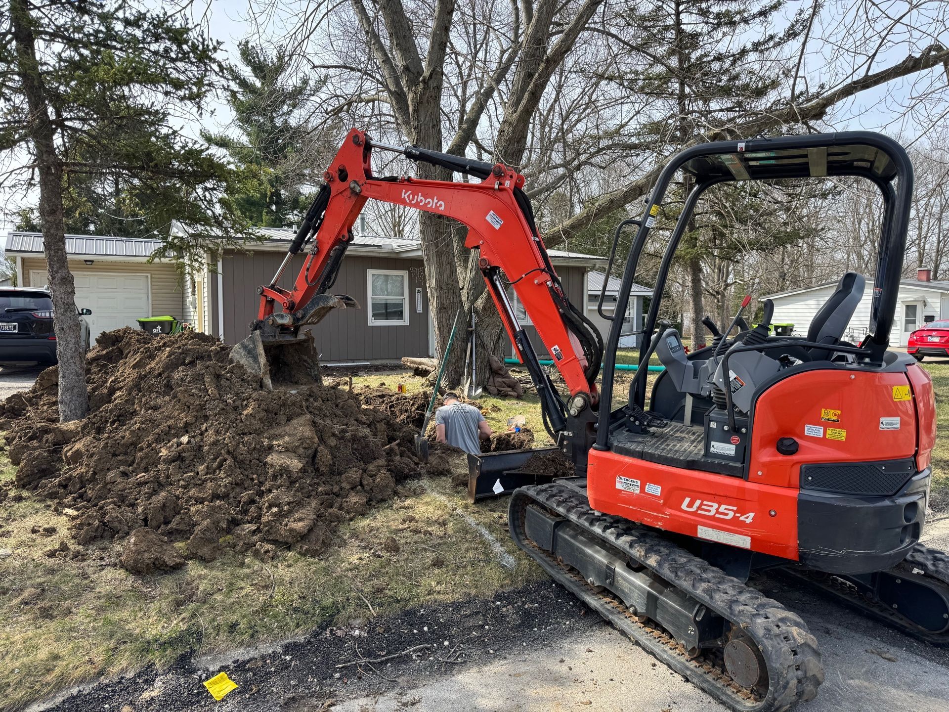 Red excavator digging a large dirt pile in a yard beside a house and trees.