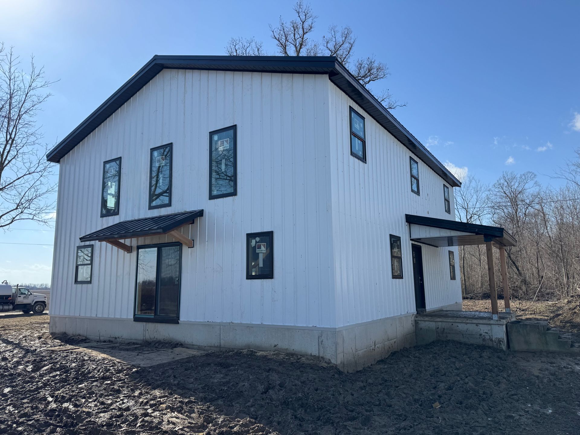 White two-story house with black trim and a metal roof on a bare lot under a blue sky.