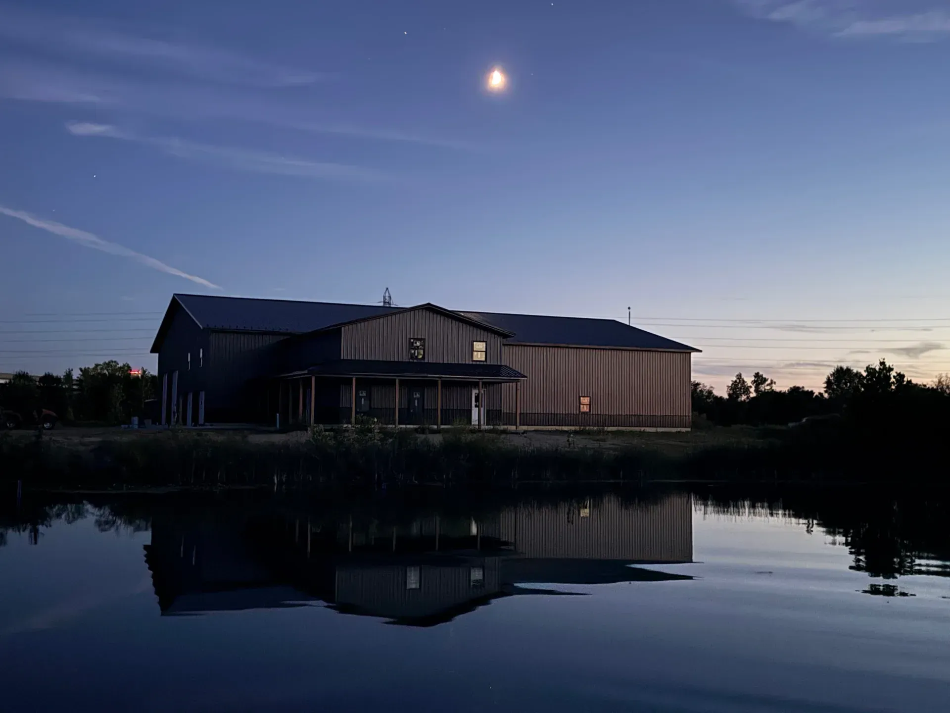 House by calm water at dusk, with a crescent moon and reflected sky.