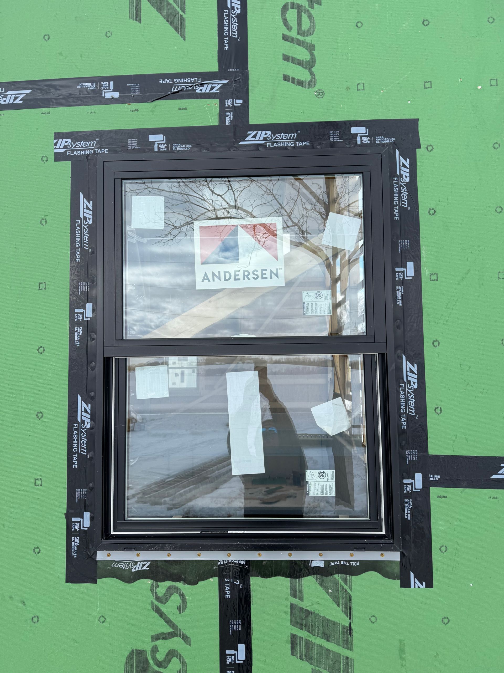 Small black-framed window set in green wall sheathing with an Andersen label and taped glass.