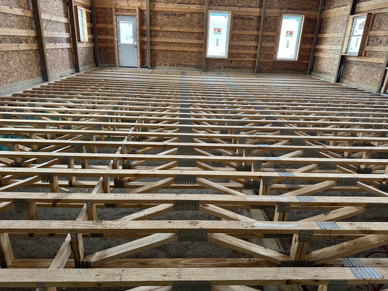 Interior construction site with exposed wooden floor joists and brick walls, viewed toward windows and a door