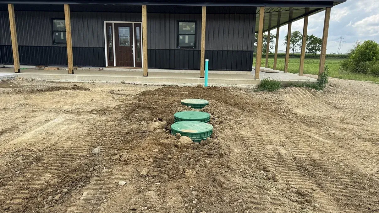 Three green utility lids in a dirt yard in front of a dark-sided building.