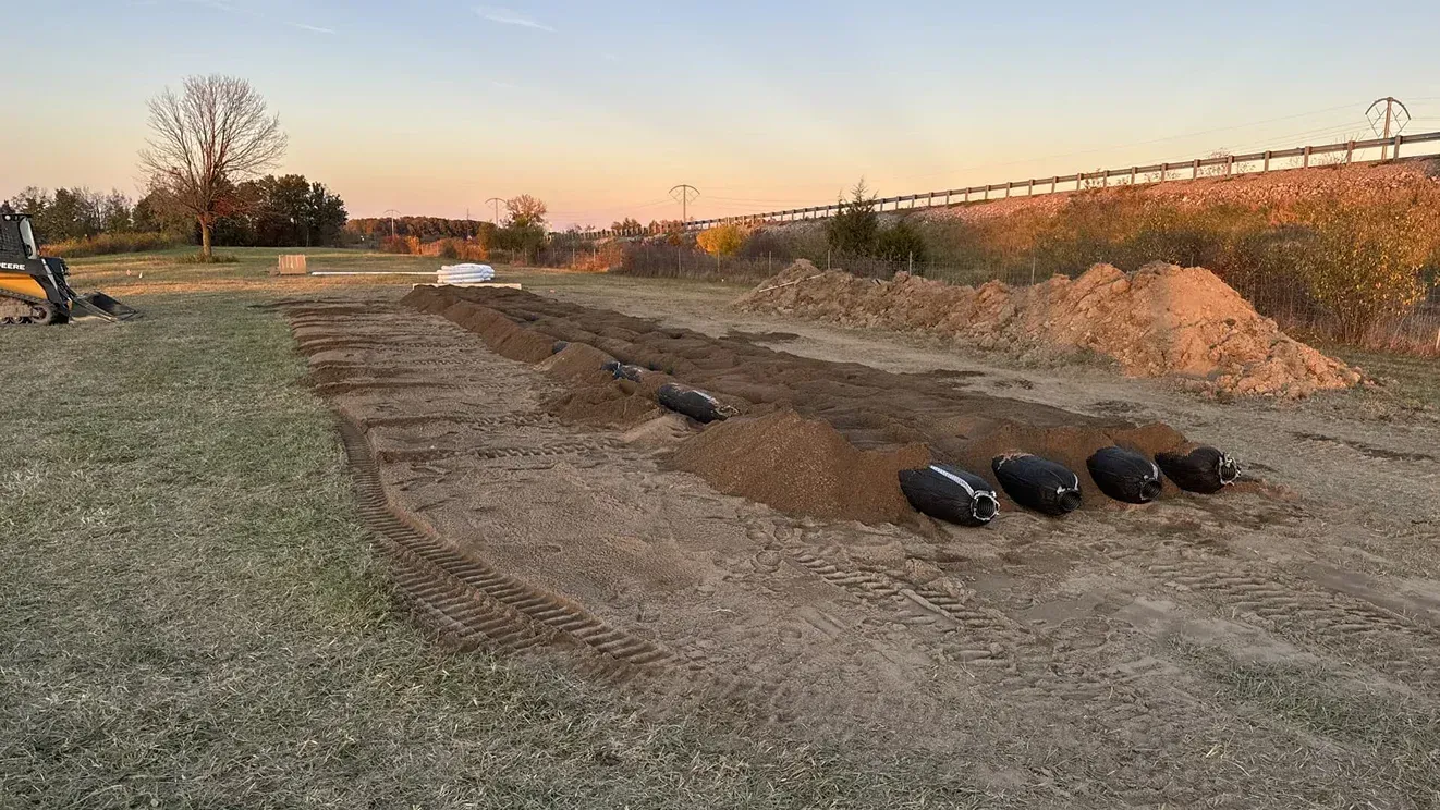 Grassy field with a curved dirt trench, black sandbags, and a small excavator at sunset