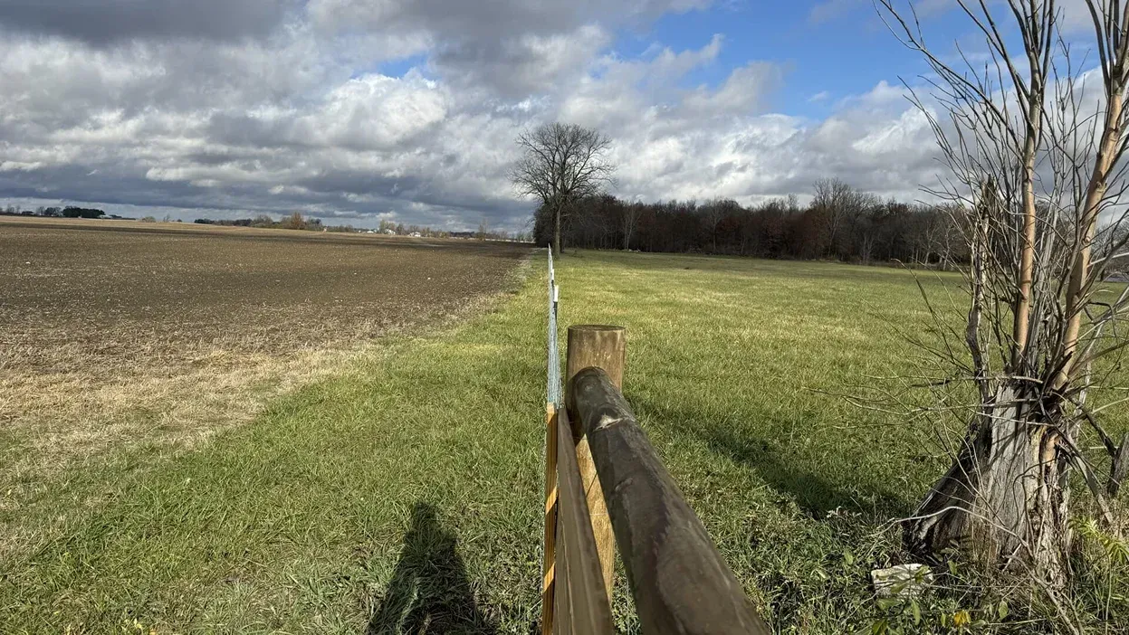 Rural field with a split fence between plowed brown land and green grass under a cloudy sky