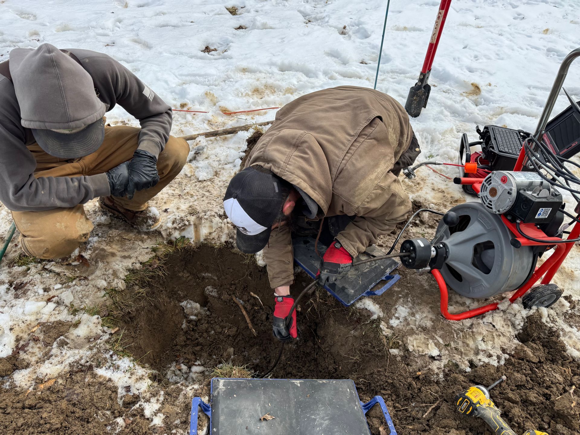 Two people digging in snowy mud beside a red auger and tools outdoors