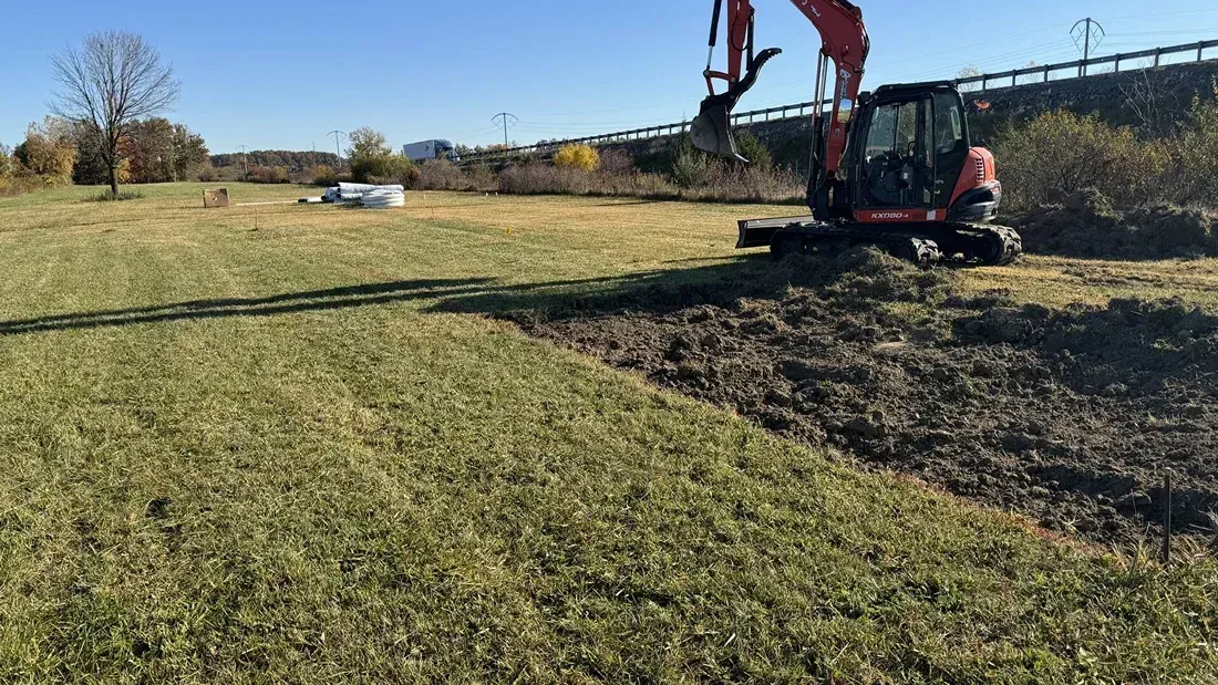 Excavator digging soil beside a grassy field under a clear sky