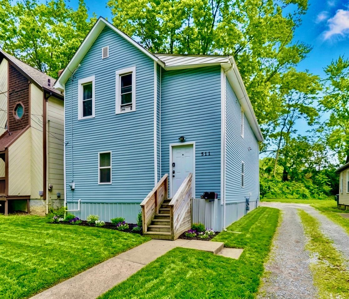 Blue house with white trim, front steps, and a gravel driveway beside a green yard and trees