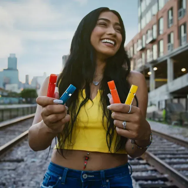 A woman in a yellow crop top and blue jeans is holding three different colored sticks in her hands.