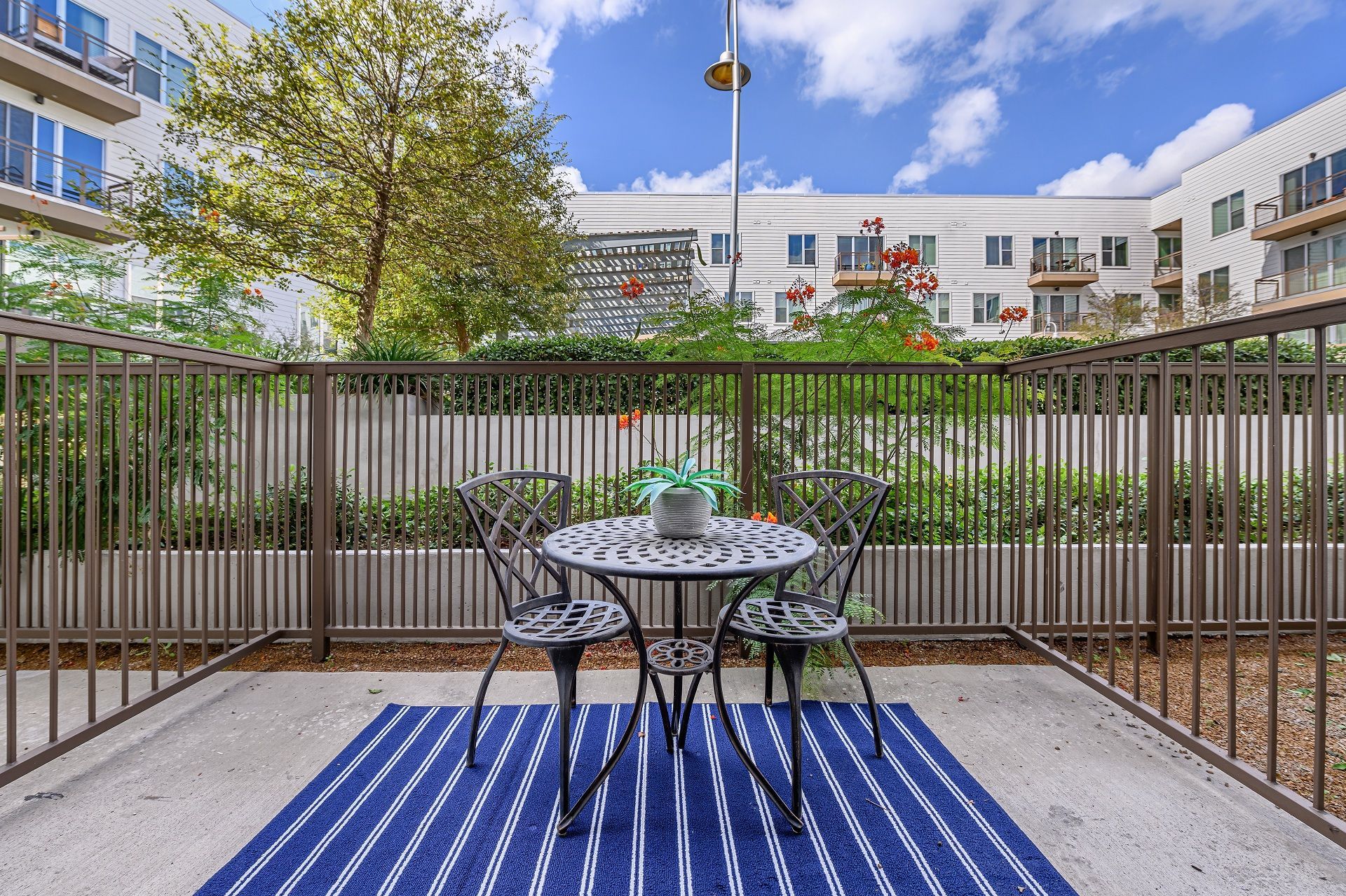 Patio with metal furniture, blue rug, and bordering fence. Apartment buildings and a bright sky are in the background at Sabina, offering affordable residences near Downtown Austin, TX.