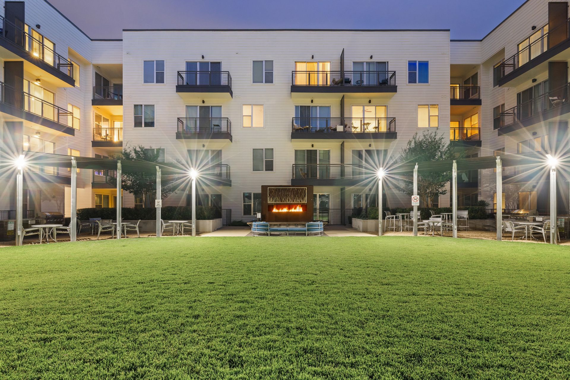 Apartment courtyard with green lawn, lit palm trees, and a modern building at dusk at Sabina in Austin, TX.