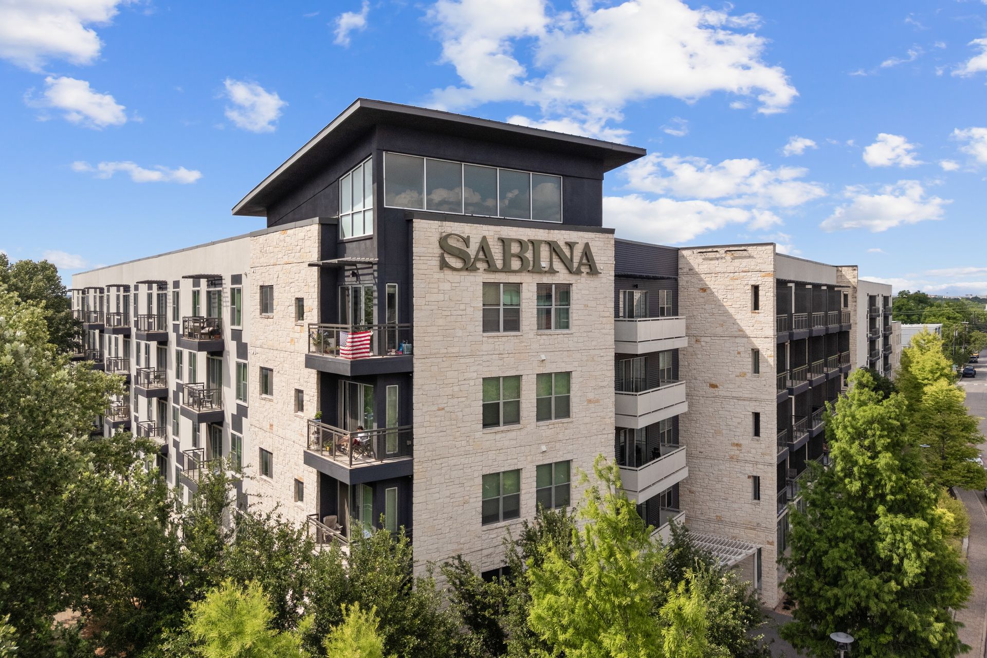 Modern apartment building with “SABINA” sign, stone facade, and rooftop terrace under a blue sky at Sabina in Austin, TX.