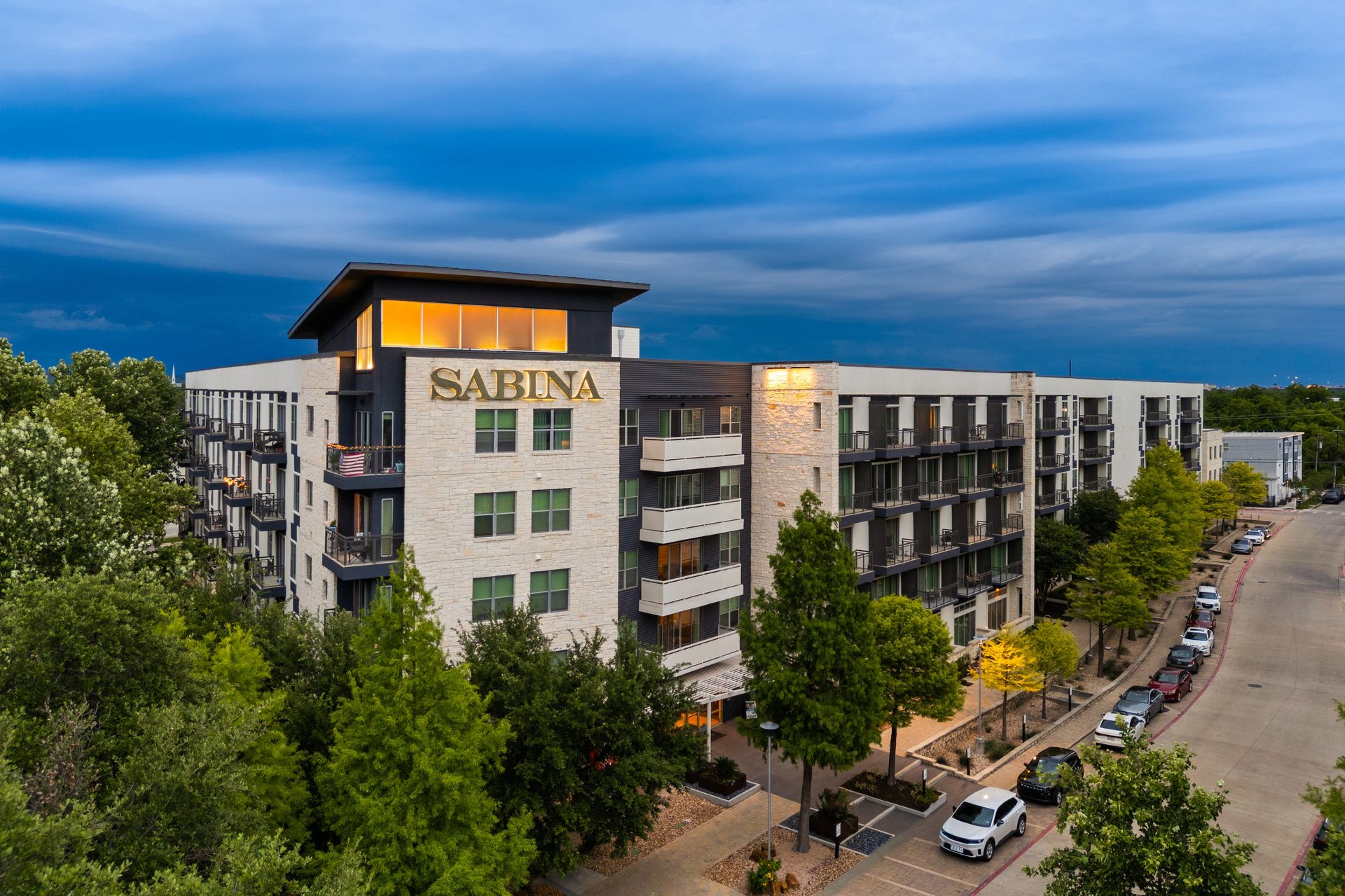 Apartment building with SABTINA sign at dusk, viewed from a tree-lined street at Sabina in Austin, TX.