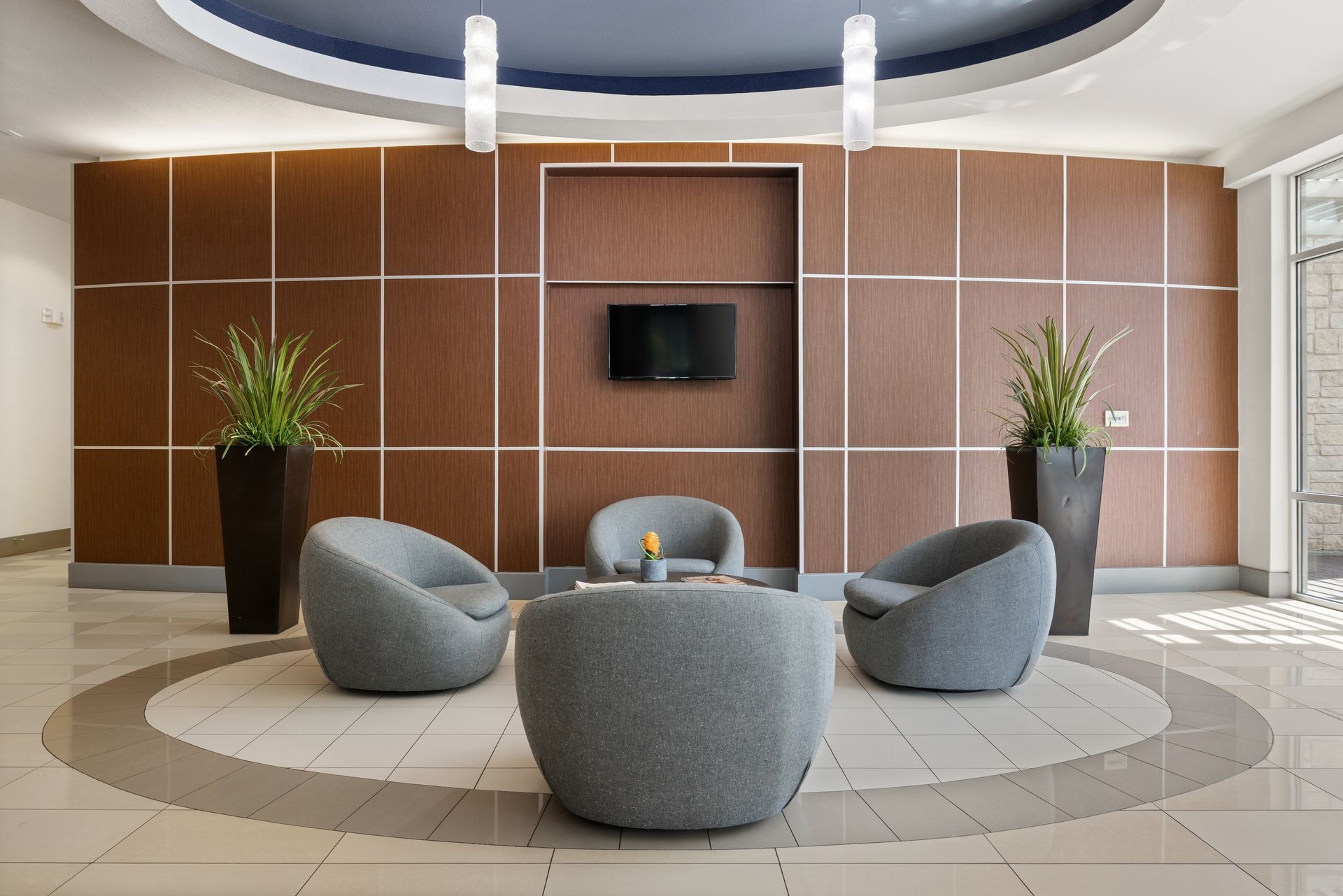 Modern lobby seating area with gray chairs, round ottoman, wood-paneled wall, and potted plants at Sabina in Austin, TX.