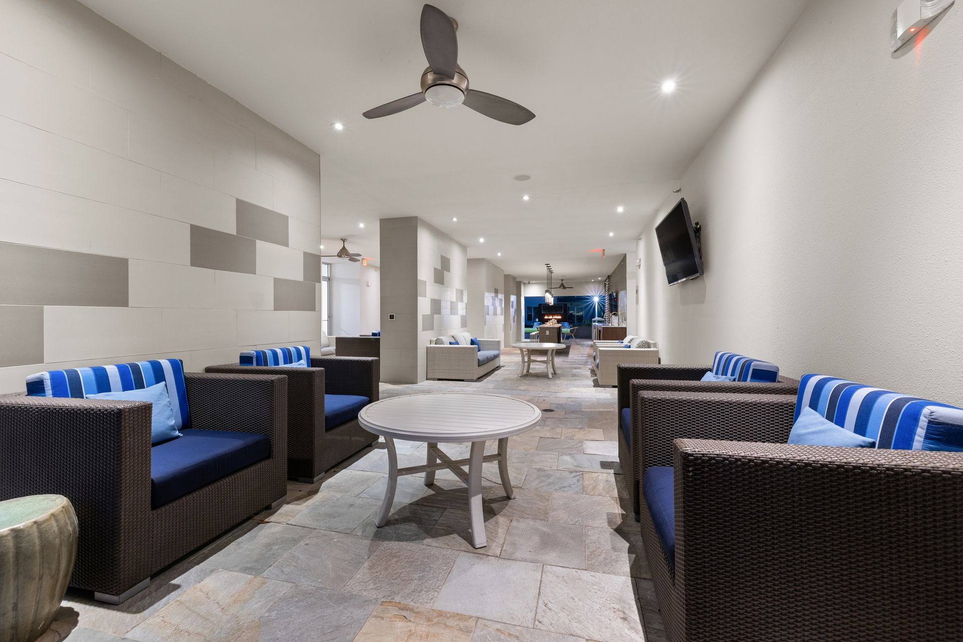 Modern lounge with wicker armchairs, blue cushions, coffee tables, ceiling fans, and a bright hallway beyond at Sabina in Austin, TX.
