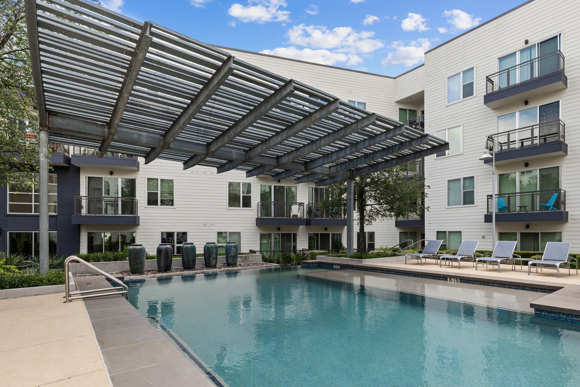 Apartment pool with a metal canopy, lounge chairs, and blue water under a partly cloudy sky at Sabina in Austin, TX.