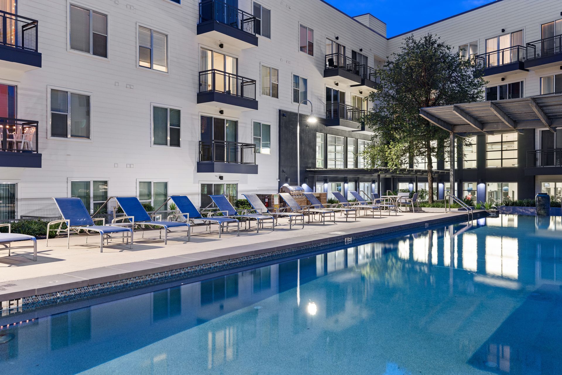 Apartment pool with blue lounge chairs and lit buildings at dusk at Sabina in Austin, TX.