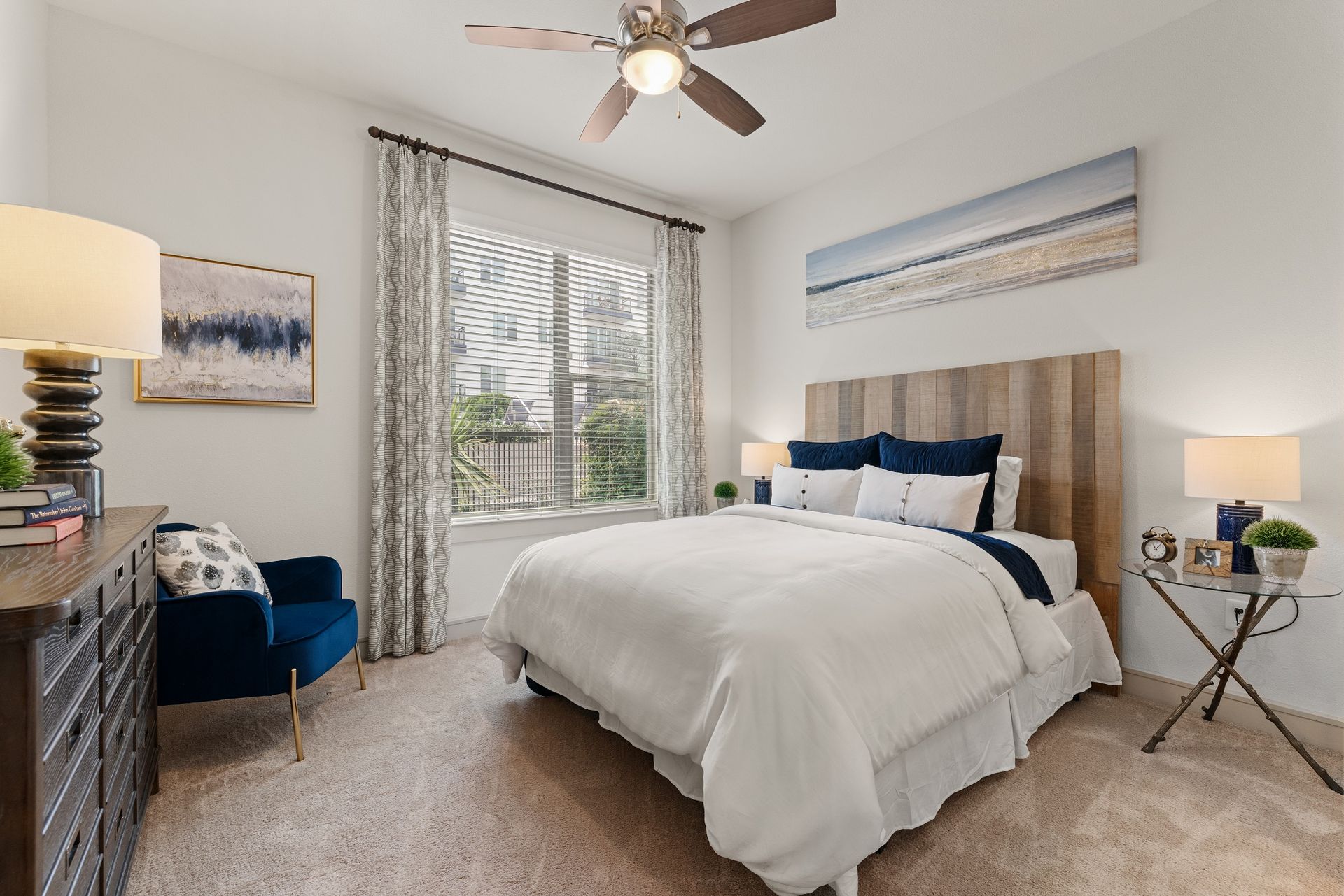 Bright bedroom with a king bed, blue accents, armchair, ceiling fan, and sliding glass doors to a balcony at Sabina in Austin, TX.