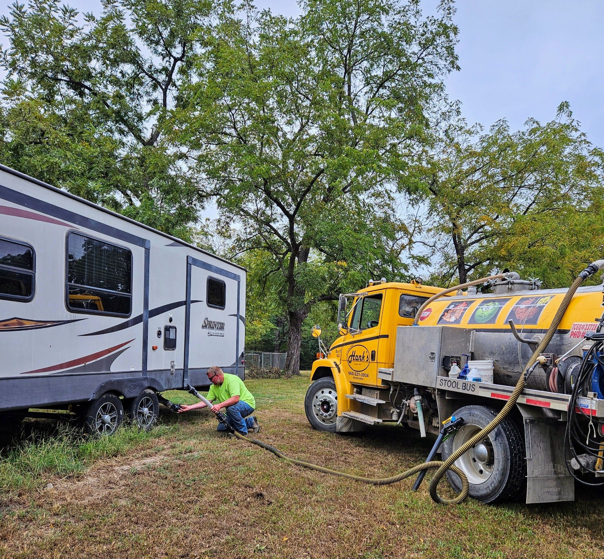 Man emptying RV waste tank with a yellow truck in a grassy area with trees.