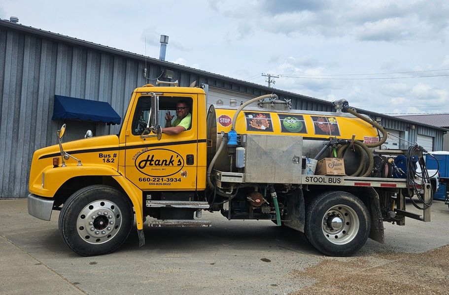 Yellow Hank's portable restroom truck with a driver in the driver's seat, parked in front of a building.