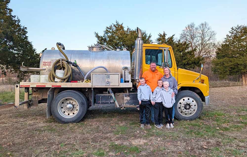 Family poses with a yellow truck and a large silver tank in a grassy field.