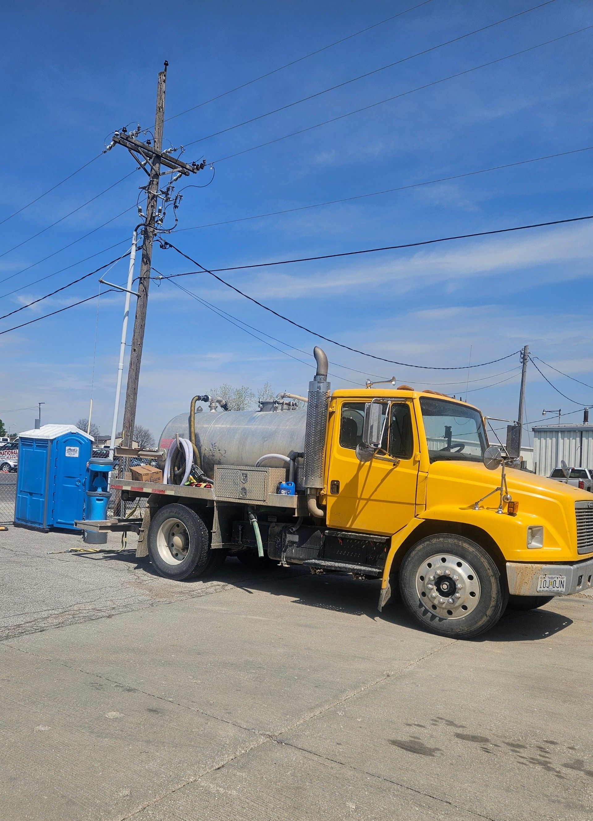 Yellow sanitation truck parked next to a utility pole and portable toilet, under a blue sky.