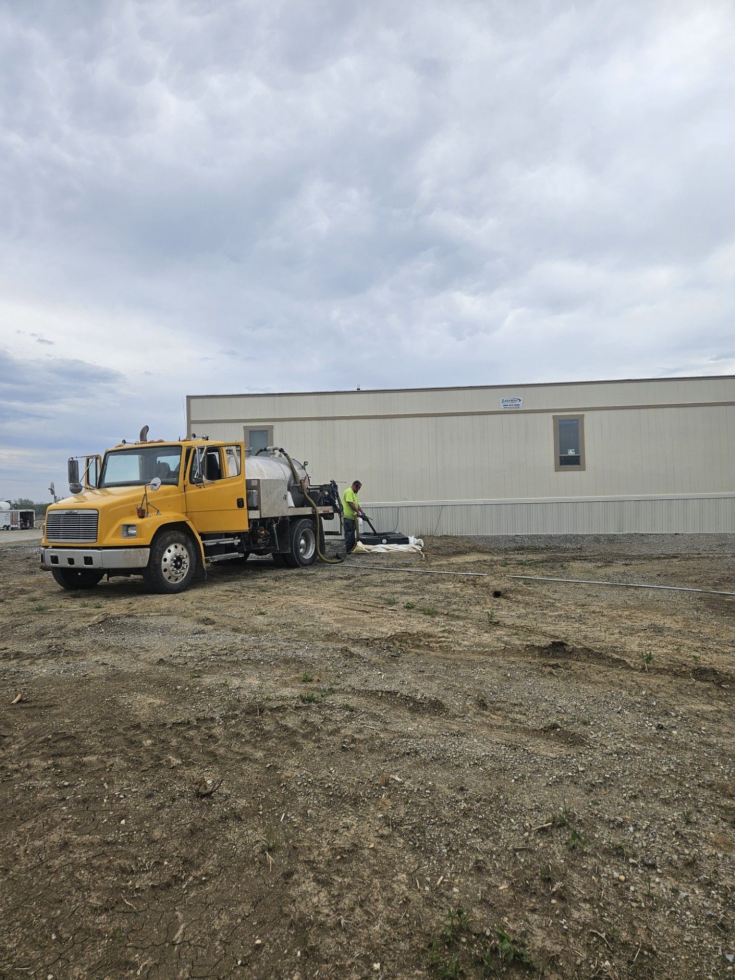 Yellow truck and worker pumping septic tank next to a mobile home on a gravel lot under cloudy skies.