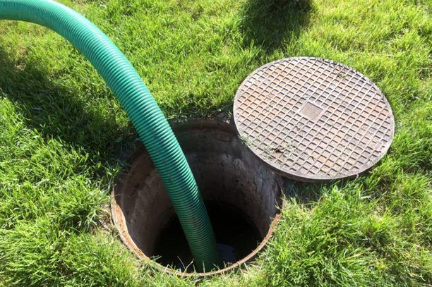 Green hose going into an open septic tank. Lid sits nearby on grass.