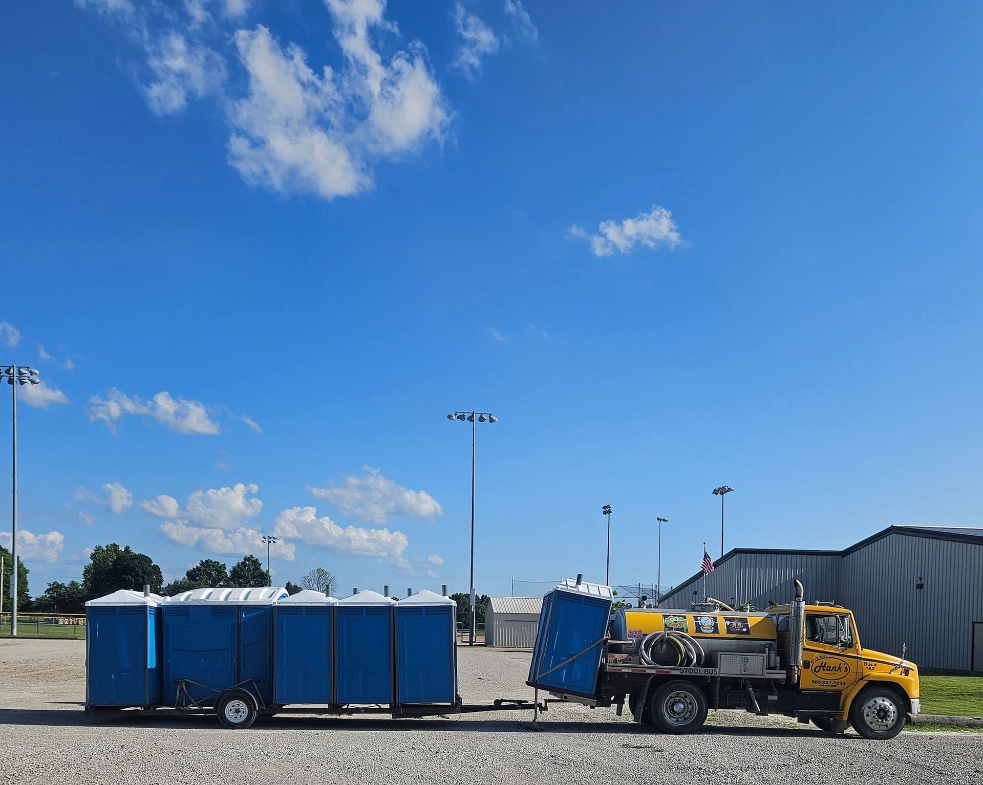 Blue portable toilets and a yellow utility truck in a gravel lot under a bright blue sky