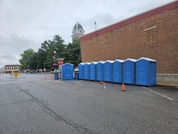 Row of blue portable toilets on a paved area; red brick building and courthouse in the background.