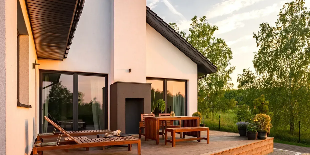 A modern house with a patio, wooden furniture, and sunbeds. Green trees are in the background.