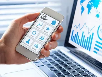 Hand holding phone displaying a real estate app, laptop with charts in background.