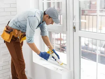 Man caulking a white window frame indoors; wearing a tool belt and gloves.